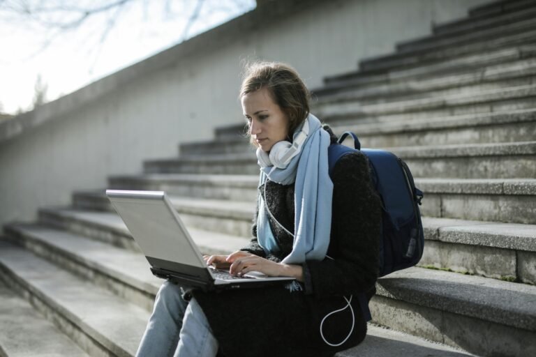 college student sitting on steps using a laptop, with a backpack on and noise cancelling earphones around her neck.
