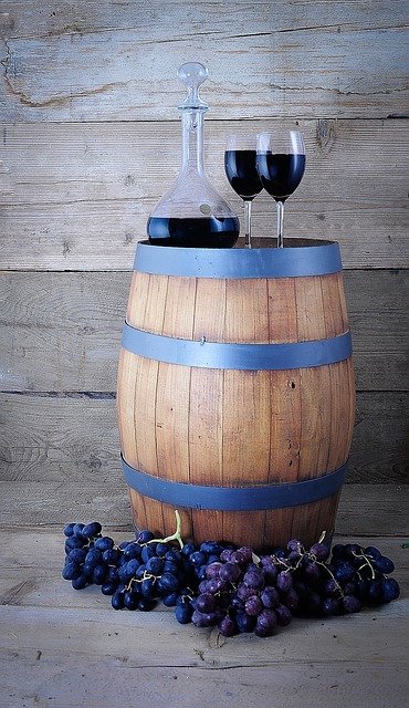 Close up photo of 2 wine glasses filled with red wine sitting on top of a wine barrel with clusters of grapes around the bottom of the barrel